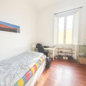 camera studio, letto a un palmo, finestra illuminata, mobili in legno, colori vivaci.