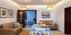 Soggiorno accogliente in appartamento vista mare con divani marroni, tavolo e lampade su pareti bianche.