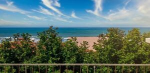 Balcone appartamento vista mare: alberi verdi, oceano blu e cielo sereno con nuvole.