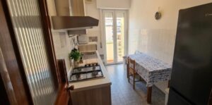 Piccola cucina accogliente in appartamento vista mare con frigo nero e tavolo blu.