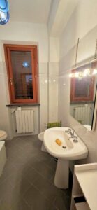 Bagno compatto con piastrelle chiare, lavabo a colonna, specchio illuminato e finestra in legno.