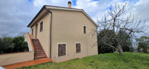 Casa di campagna beige con scala esterna, persiane in legno e giardino.