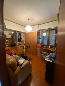 Stanza disordinata con poltrona beige, bici statica e scrivania alla luce soffusa.