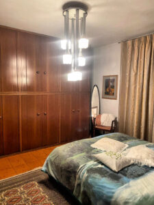 Camera da letto con lampadario, armadio scuro, letto blu verde e poltrona arancio.