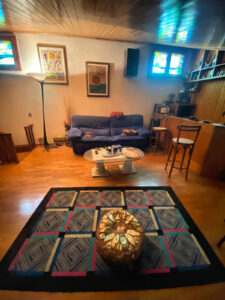 Soggiorno accogliente con tappeto geometrico, pouf tondo e divano blu su parquet in legno.