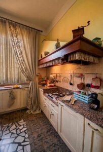 Angolo cucina giallo con mensole legno, lavello granito, tende crema e pavimento mosaico marrone.