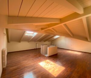 Soffitta vuota luminosa con tre lucernari, travi in legno e parquet scuro.