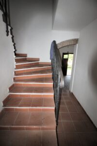 Scala interna in cotto con corrimano metallico che conduce a porta luminosa verso giardino verde