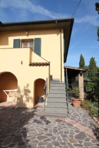 Cortile casa mediterranea gialla con scala esterna in pietra, pavimento irregolare e giardino soleggiato