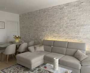 Soggiorno moderno con sofa L morbida, parete texturizzata grigio-beige e tavolino decorato.