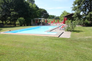 Piscina all’aperto con acqua azzurra, prato verde e lettini rossi ombreggiati da alberi
