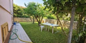 Prato verde con tavolo bianco, graticcio e casa rosa; tubo e annaffiatoio blu nel giardino.