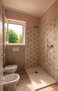 Bagno rosa con doccia fiorata, wc e finestra su alberi: luminoso ed elegante.