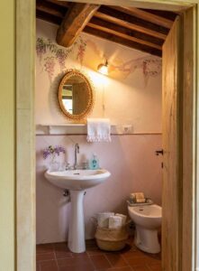 Bagno rustico italiano con lavabo a colonna, bidet, travi in legno, pavimento in cotto e fiori viola