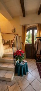Ingresso rustico con scala, porta in legno, fiori rossi, travi a vista e pavimento piastrellato.