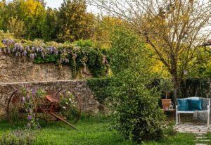 Tenuta con fonte romana, giardino rustico con panchina bianca, glicine e attrezzo agricolo.