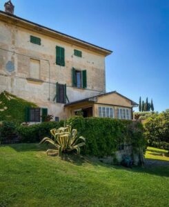 Casa rustica in pietra con giardino curato, siepi, succulenta e piccola serra sotto cielo sereno.