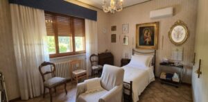 Accogliente camera beige con letto singolo, poltrona bianca e due sedie vicino alla finestra luminosa.