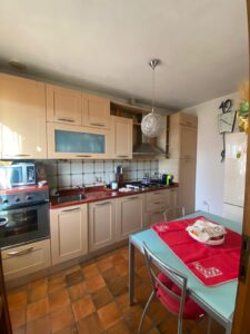 Cucina piccola luminosa con top rosso, frigo bianco e tavolo apparecchiato su pavimento marrone.