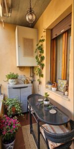 Terrazza soleggata con tavolo nero, sedie rattan e piantane verdi.