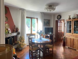 Sala da pranzo con tavolo rotondo, sedie imbottite, caminetto rosso e luce naturale dalla finestra.