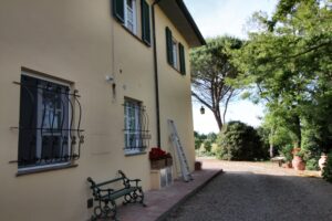 Villa gialla con persiane verdi e viale di ghiaia tra alberi, tranquilla in campagna.
