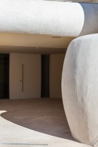 Ingresso architettonico moderno con forme organiche in cemento e cortile esterno luminoso.
