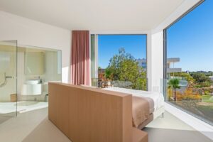 Camera dalbergo minimalista con letto in legno, bagno a vista e grandi vetrate luminose.