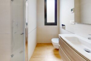 Bagno moderno e stretto con doccia in vetro, sanitari bianchi e mobile in legno.